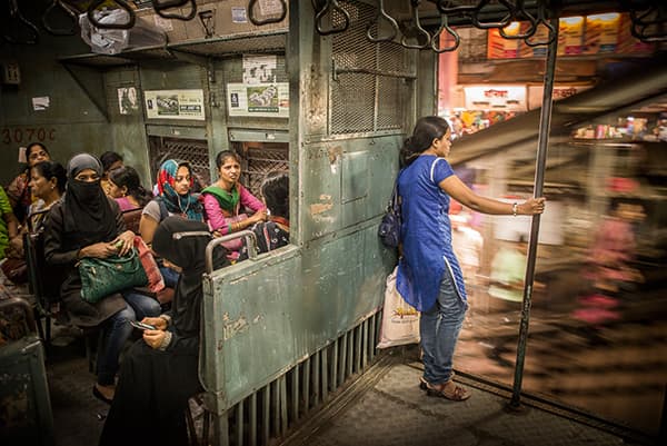 A woman looking into the Mumbai night. Beside her we see women from a variety of Indian cultures, all of whom sit in the ladies’ compartment of this local train