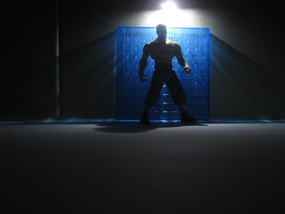 Silueta de un muñeco luchador de juguete frente a un panel azul con una lampara alumbrando desde encima de su cabeza.